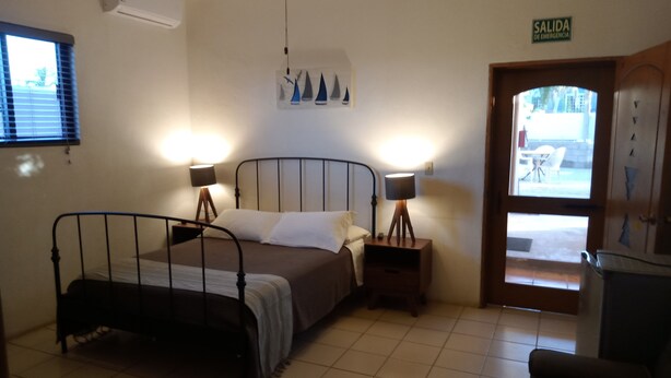 Un dormitorio con cama, dos mesitas de noche con lámparas, una ventana con persianas, una puerta con un cartel y piso de baldosas.