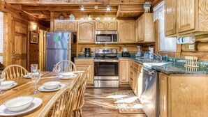 Une cuisine rustique aménagée dans une cabane en bois, avec un coin repas, des meubles en bois, des appareils électroménagers en acier inoxydable et une fenêtre équipée de stores.