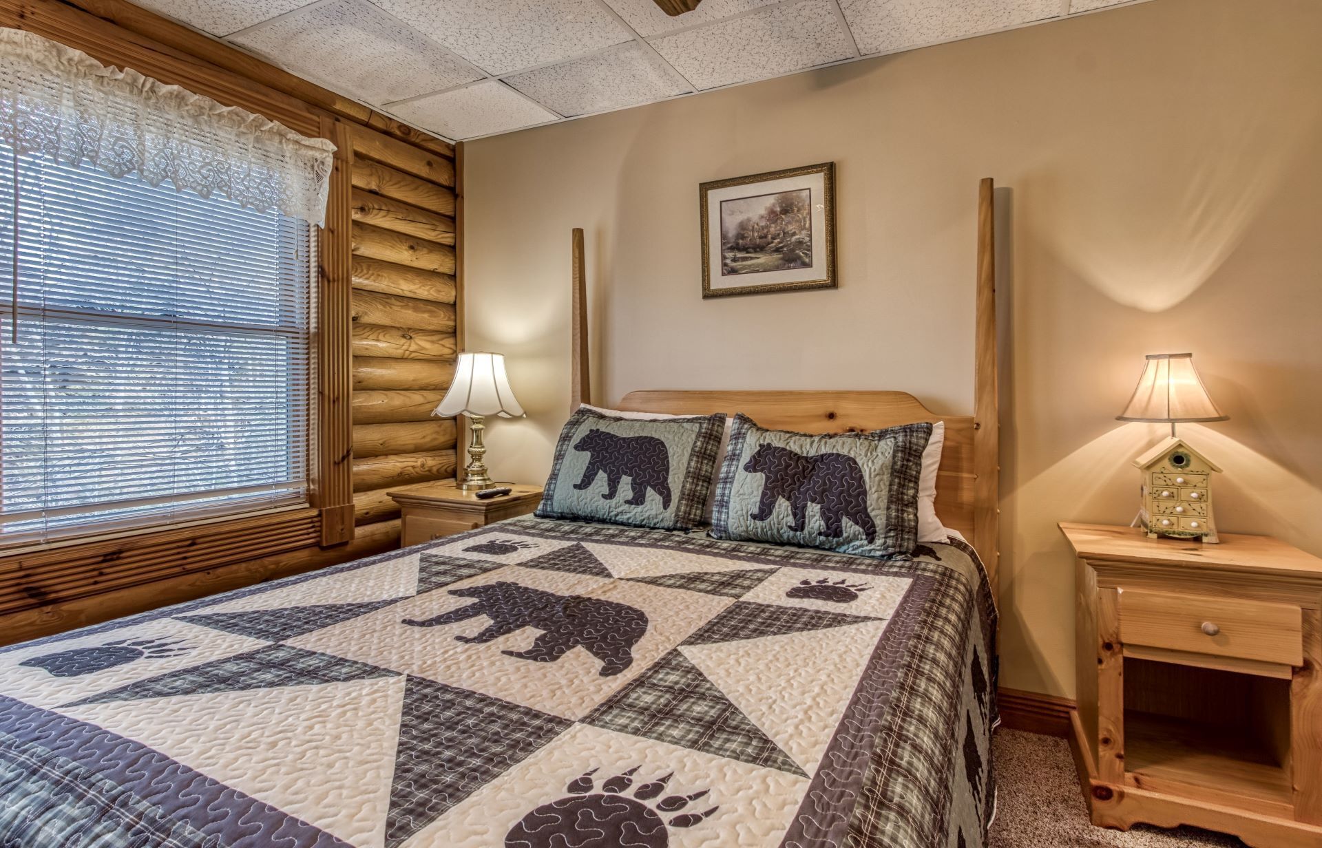 A cozy bedroom with a wooden headboard, a quilted bedspread featuring animal paw prints, a bedside lamp, and a small wooden nightstand.