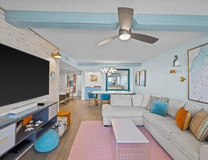 A modern living room with a flat-screen TV, a ceiling fan, and a dining area with blue chairs.