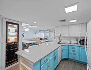 A modern kitchen with blue cabinets, white countertops, and a central island. There is a dining area with a sofa and a coffee machine.