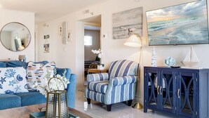 A living room with a blue sofa, striped armchair, wooden coffee table, and a cabinet with decorative items. - 🌅 Islander #201 – Sunrise Views & Steps to Destin’s Emerald Waters! 🌴 (Destin)