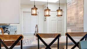 A modern kitchen with a white countertop, a sink, and a window with blinds. There are three wooden chairs with black metal frames and white cushions. - 🌅 Islander #201 – Sunrise Views & Steps to Destin’s Emerald Waters! 🌴 (Destin)