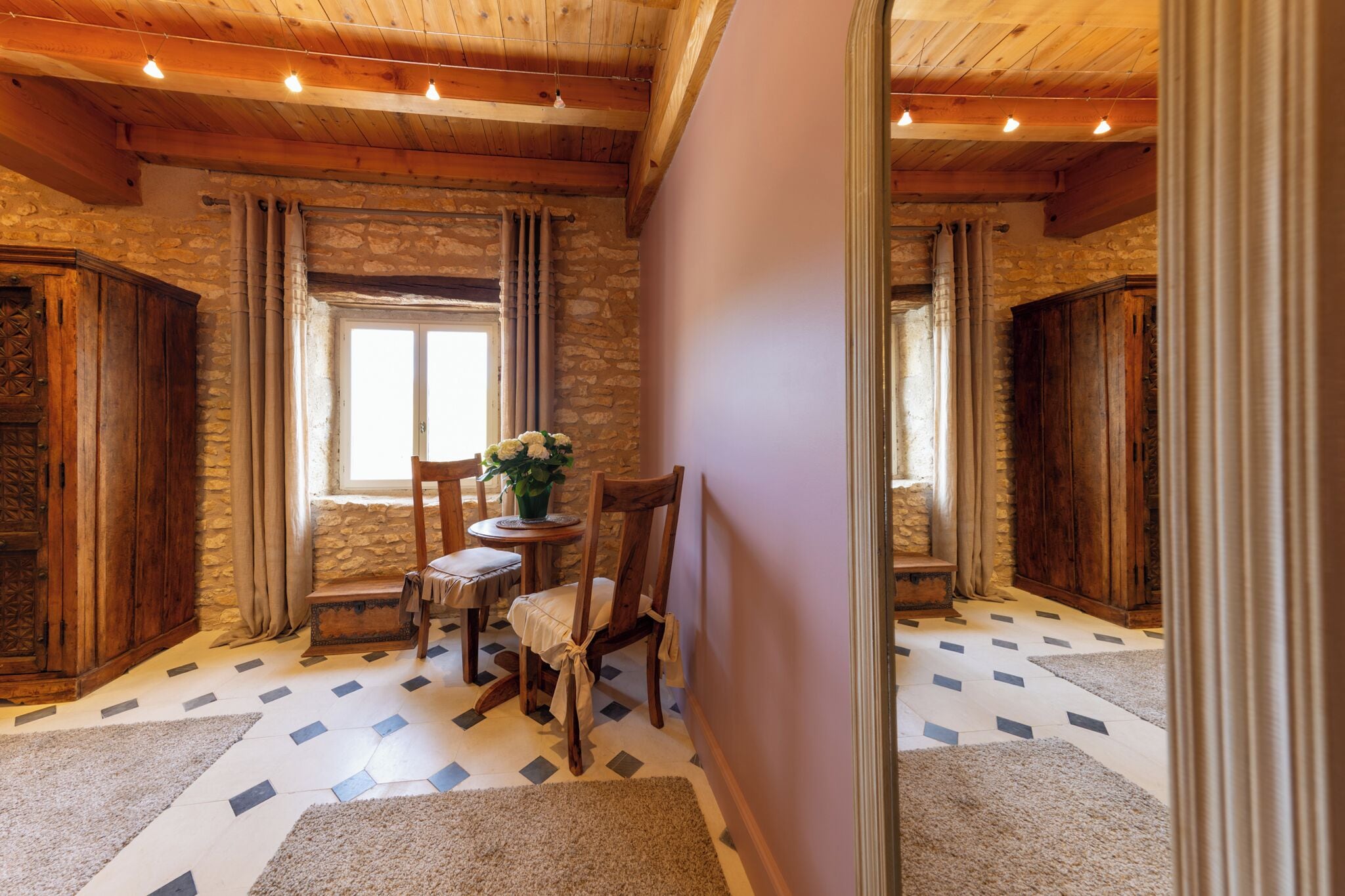 A cozy room with stone walls, wooden beams, a stone fireplace, a dining table with chairs, and a window with curtains.