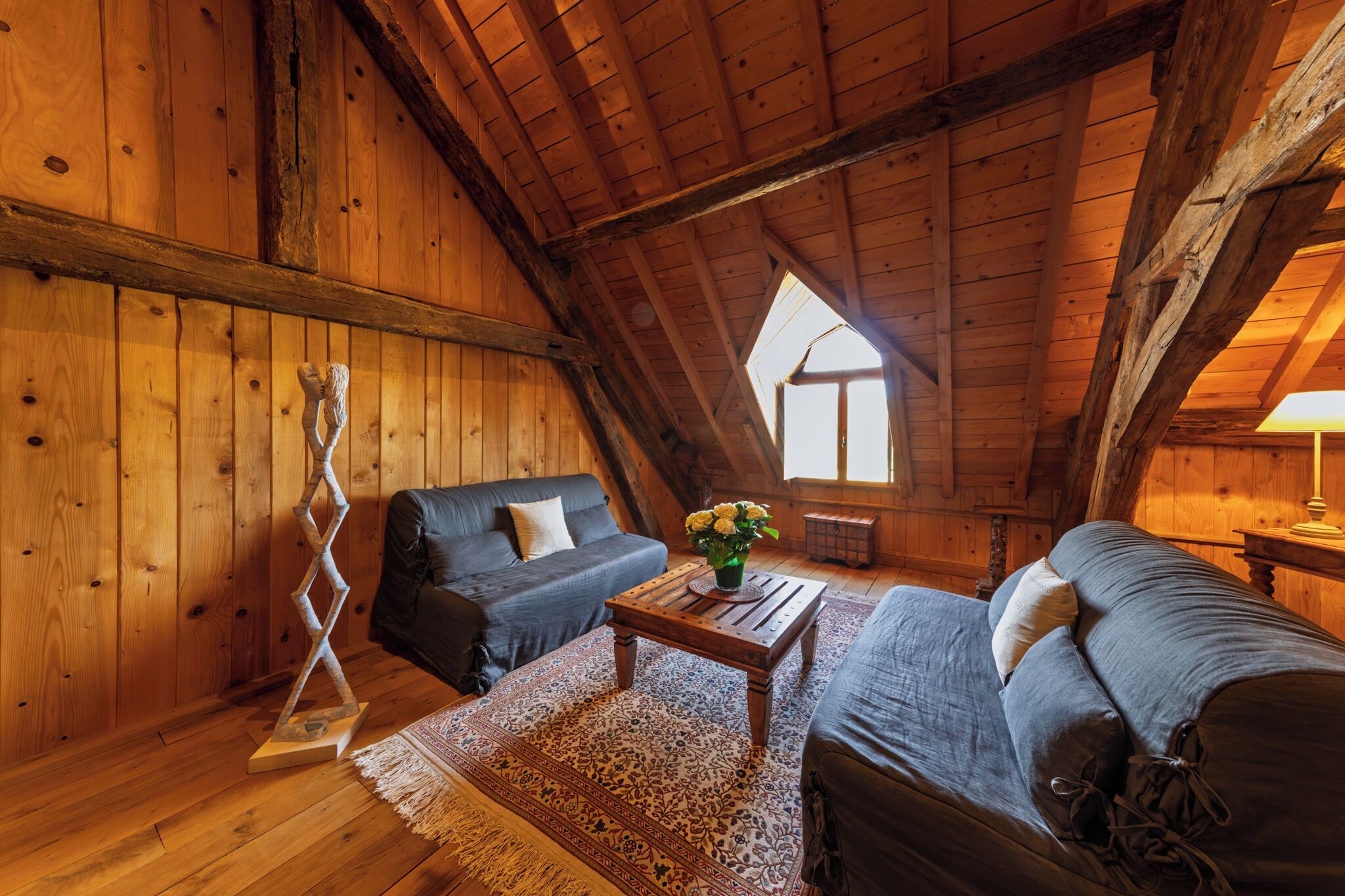 Une pièce mansardée chaleureuse aux murs en bois et au plafond en pente, comprenant deux canapés sombres, une table basse en bois et une sculpture décorative.