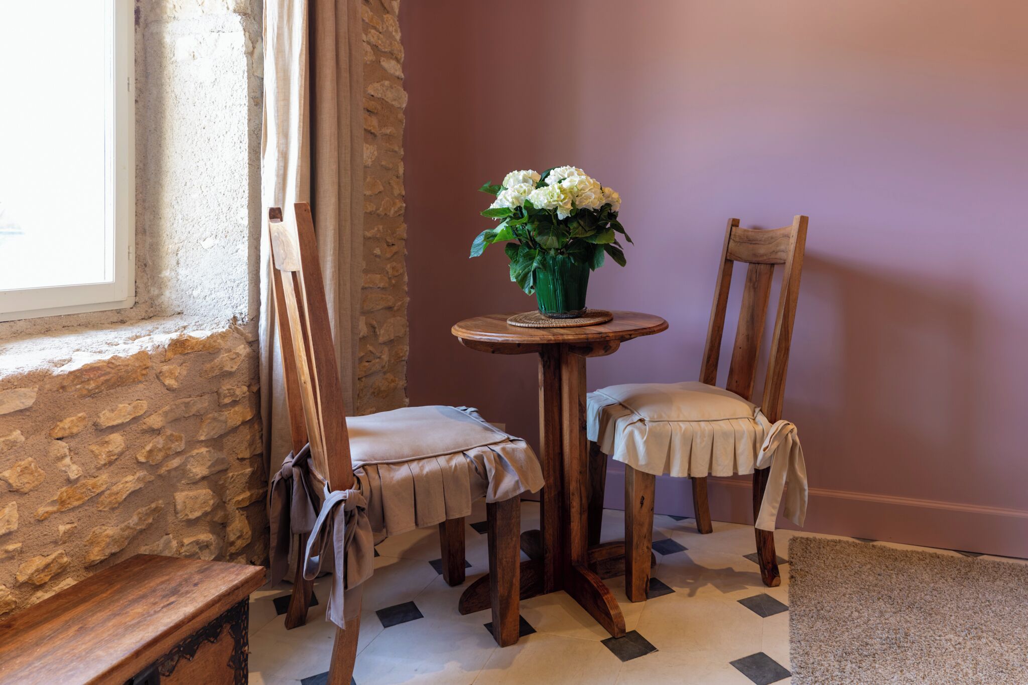 A room with a stone wall, a wooden table with a vase of flowers, two chairs, and a bench.