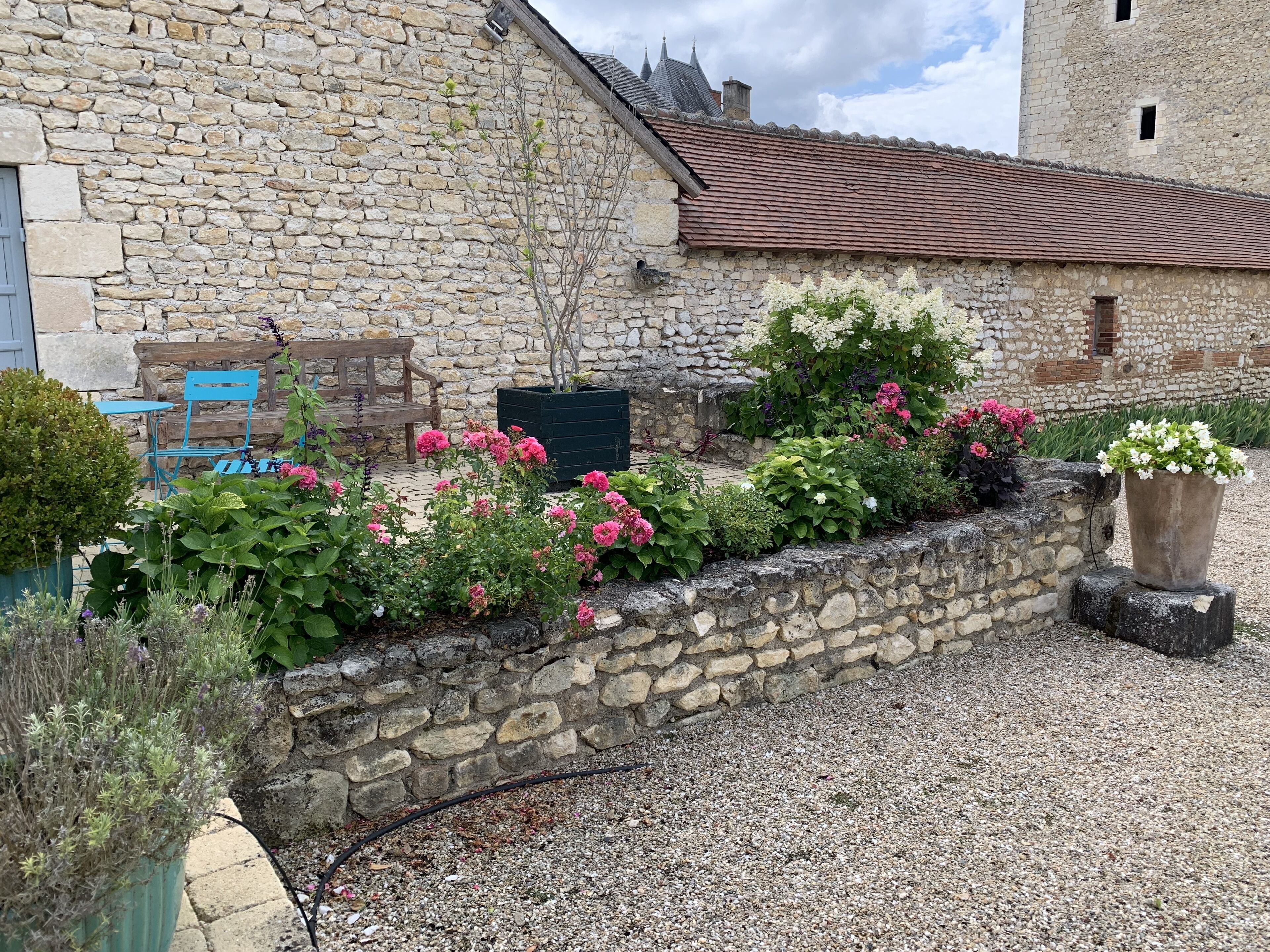 A stone building with a gravel driveway, a wooden bench, and a variety of flowering plants.