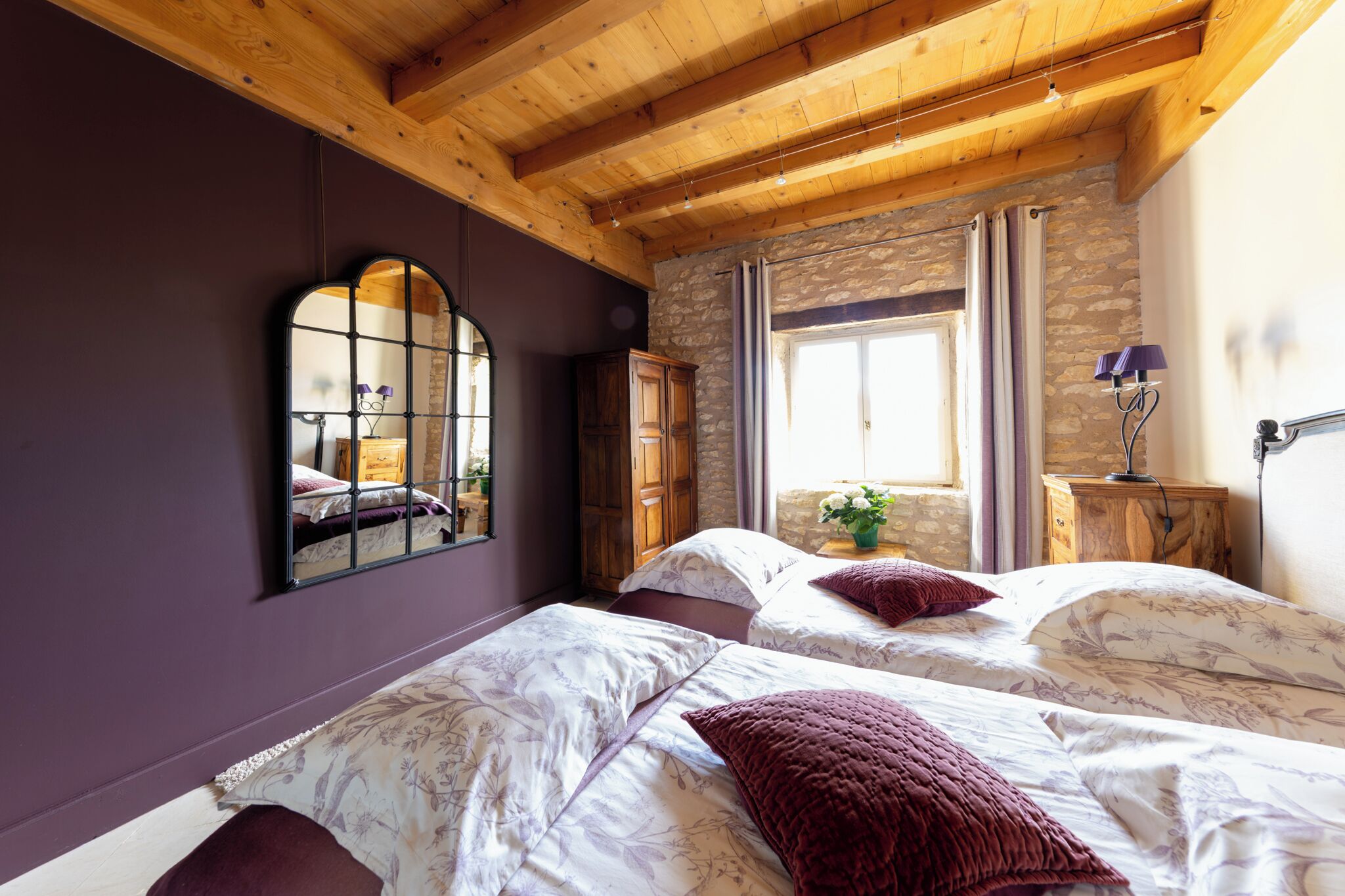 Un dormitorio con dos camas, vigas de madera, paredes de piedra, un espejo y una ventana con cortinas.