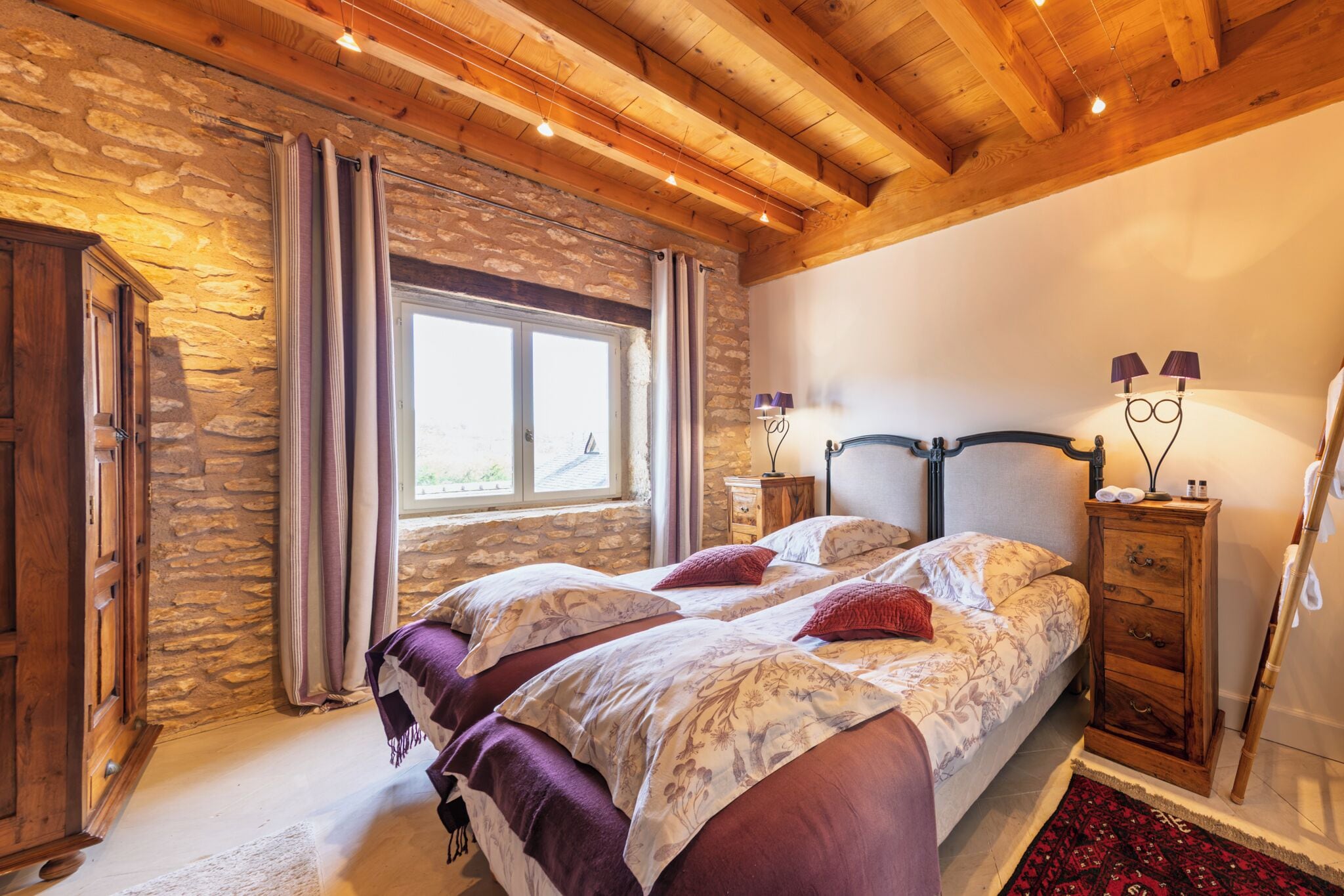 Een slaapkamer met een stenen muur, houten balken in het plafond, een bed met bloemenbeddels, twee nachtkastjes met lampen en een raam met gordijnen.