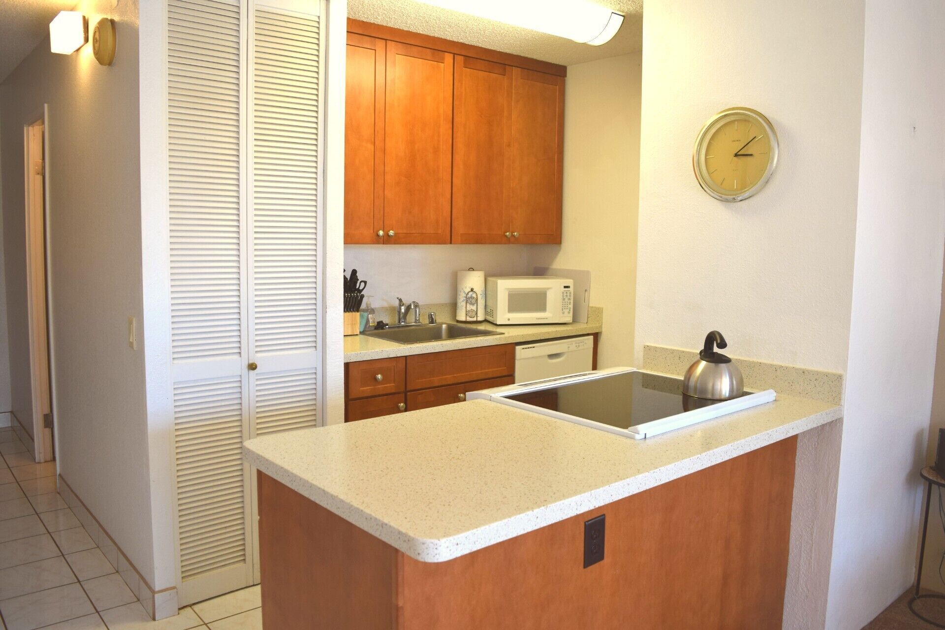 A compact kitchen with wooden cabinets, a microwave, a kettle, and a clock on the wall.