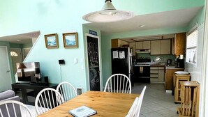 A dining area with a wooden table and white chairs, a kitchen in the background, and a sign on the wall.