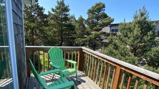 A wooden deck with two green Adirondack chairs, surrounded by a wooden railing and dense trees in the background.