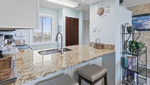 A kitchen with a granite countertop, stainless steel sink, and a built-in oven. There are wicker baskets and a metal shelf with plants. - Coastal Comfort & Resort Fun – Islander 613 (Destin)