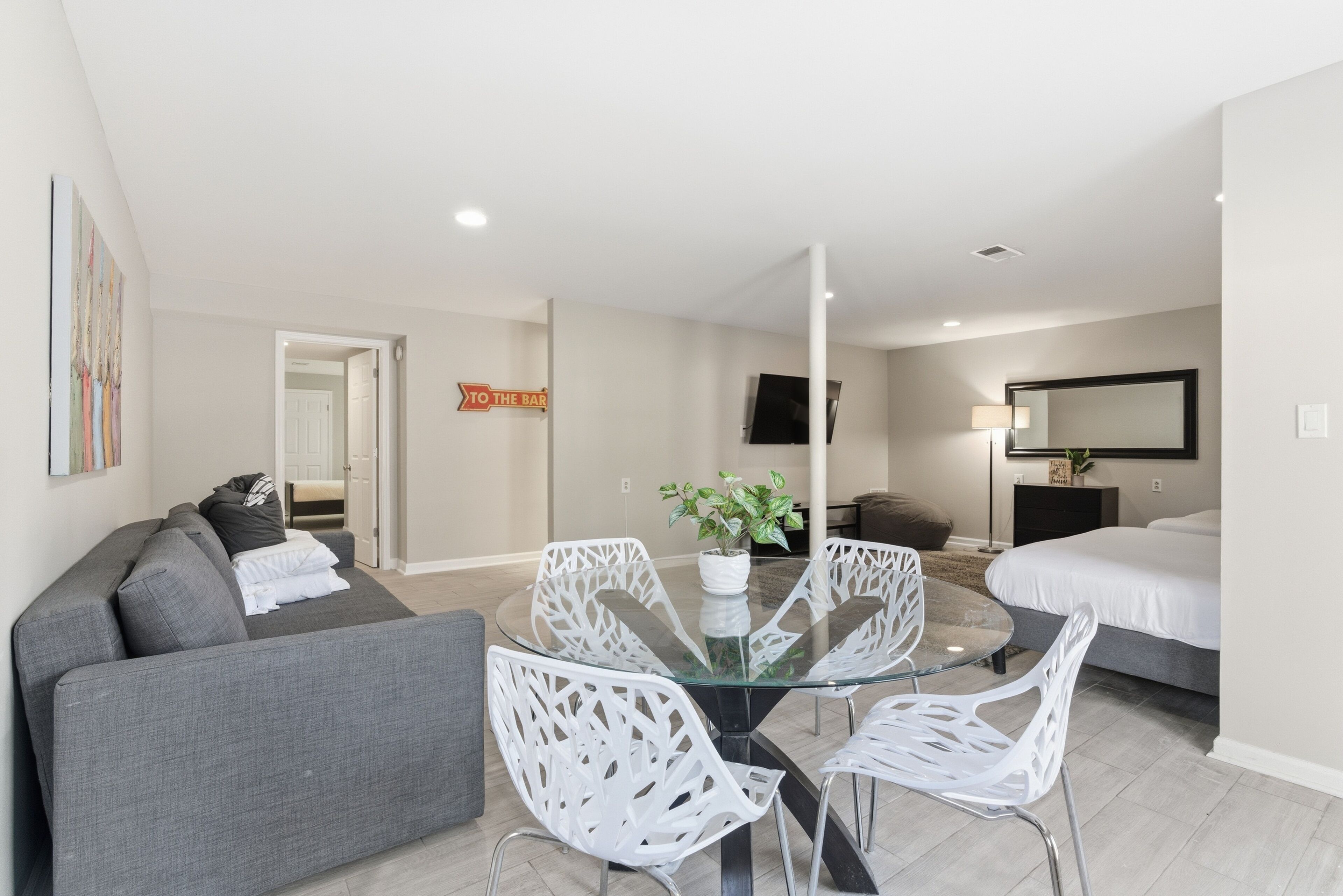 A modern hotel room with a glass dining table, white chairs, a grey sofa, a bed, a TV, and a 'TO THE BAR' sign.