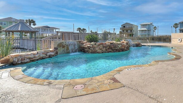 A swimming pool with a waterfall feature, surrounded by a stone wall and a wooden deck.