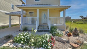 A two-story house with a front porch, a small garden with flowers, and a clear sky.