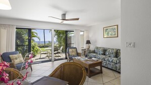 Ein großzügiges Wohnzimmer mit einer großen Glasschiebetür, die auf eine Terrasse führt, einem Deckenventilator und einem tropisch gestalteten Sofa.