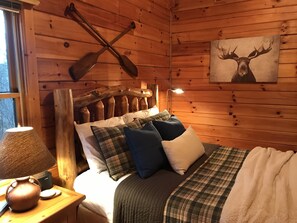 A cozy cabin room with a wooden headboard, a bed with plaid bedding, a lamp, and a painting of a moose on the wall.