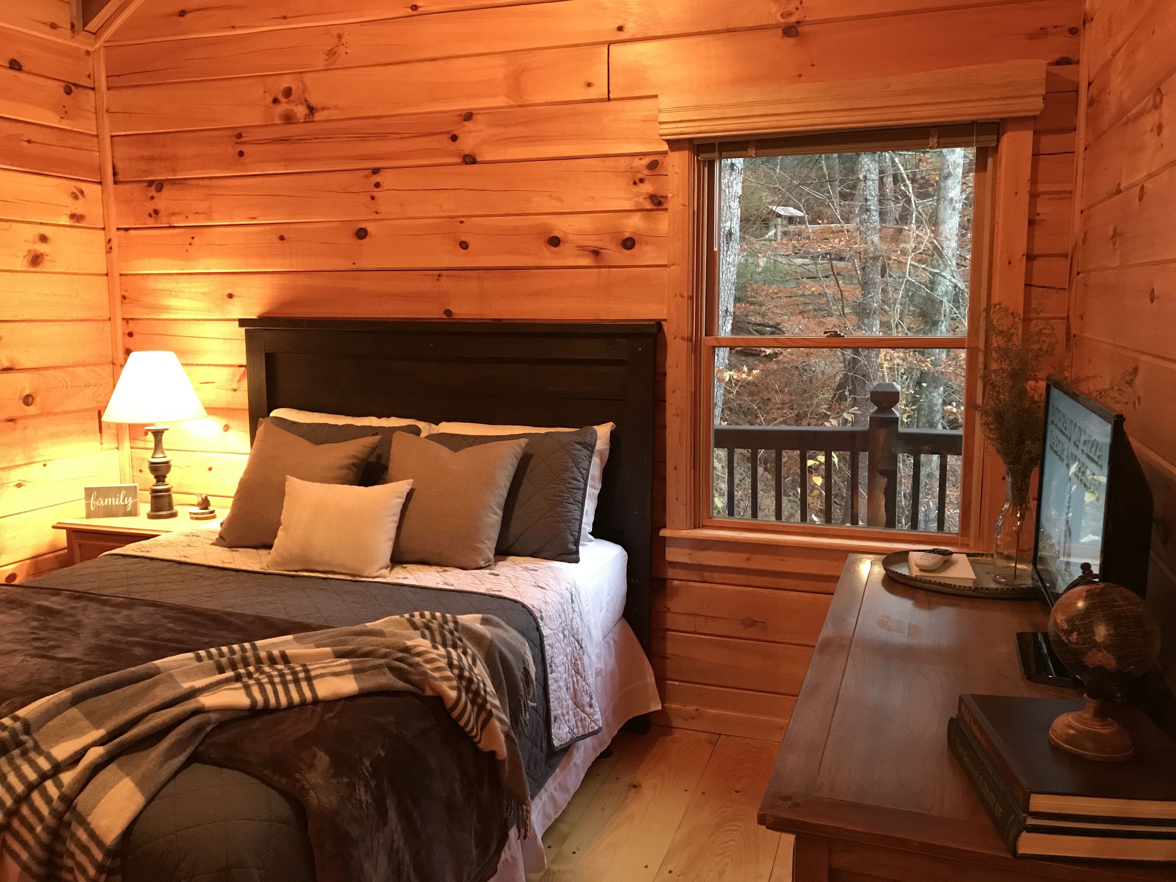 Un dormitorio de cabaña de madera acogedor, con una cama, una mesita de noche con una lámpara, una ventana con vistas a árboles y un escritorio con un globo terráqueo y libros.