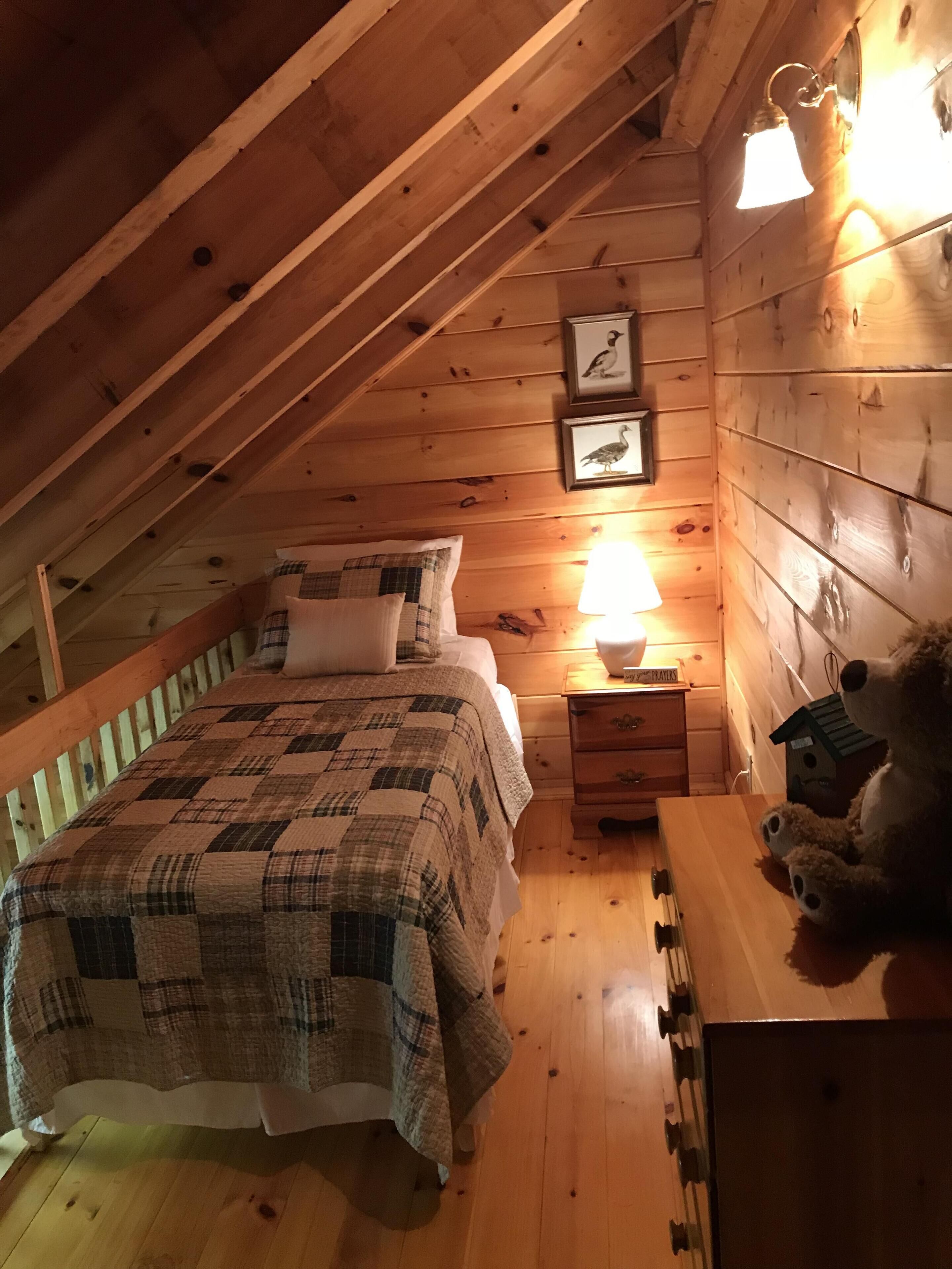 Une chambre mansardée chaleureuse avec un plafond en pente en bois, un lit recouvert d’une couette à carreaux, une table de chevet avec une lampe et un ours en peluche posé sur une table.