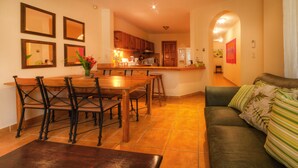 A spacious living area with a wooden dining table and chairs, a green sofa with patterned cushions, and a kitchen in the background. - Heaven On Earth! Perfect Get-Away! Gorgeous Beach. Pool. Couples &/or Families. (Samara)