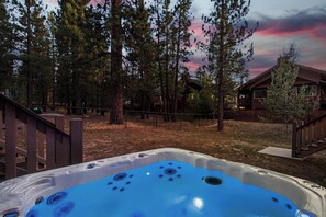 Un jacuzzi en un área boscosa con una terraza de madera y una casa al fondo.