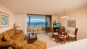 Habitación de hotel con sofá, mesa de comedor, sillas, televisor y balcón con vista al océano.