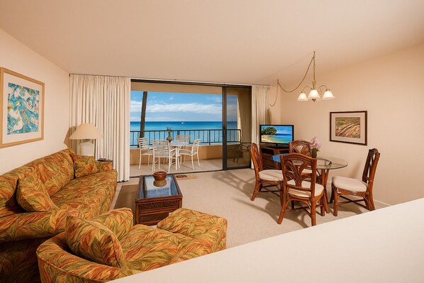 A hotel room with a sofa, dining table, chairs, a TV, and a balcony with a view of the ocean.