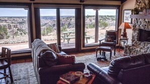 A living room with a leather sofa, a wooden coffee table, and a stone fireplace. Large windows offer a view of a rocky landscape. - Maverick cabin North Rim (Canyon)