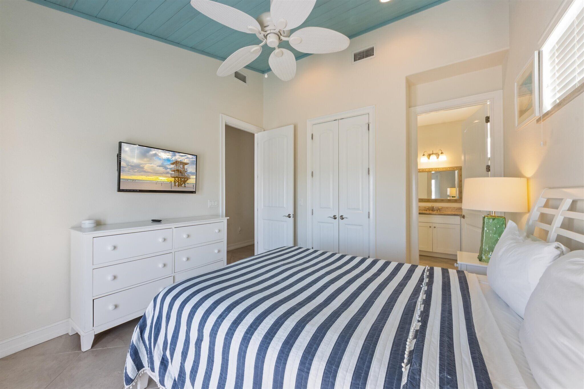Un dormitorio con cama, cómoda, ventilador de techo y baño visible a través de una puerta abierta.