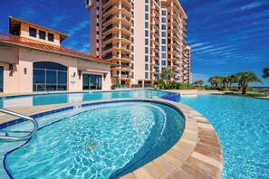 Una piscina con borde curvo, un edificio con varios balcones y un cielo azul despejado.