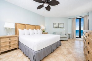 Habitación de hotel con una cama grande, ventilador de techo, mesita de noche con lámpara, sofá, cómoda y vista al océano.