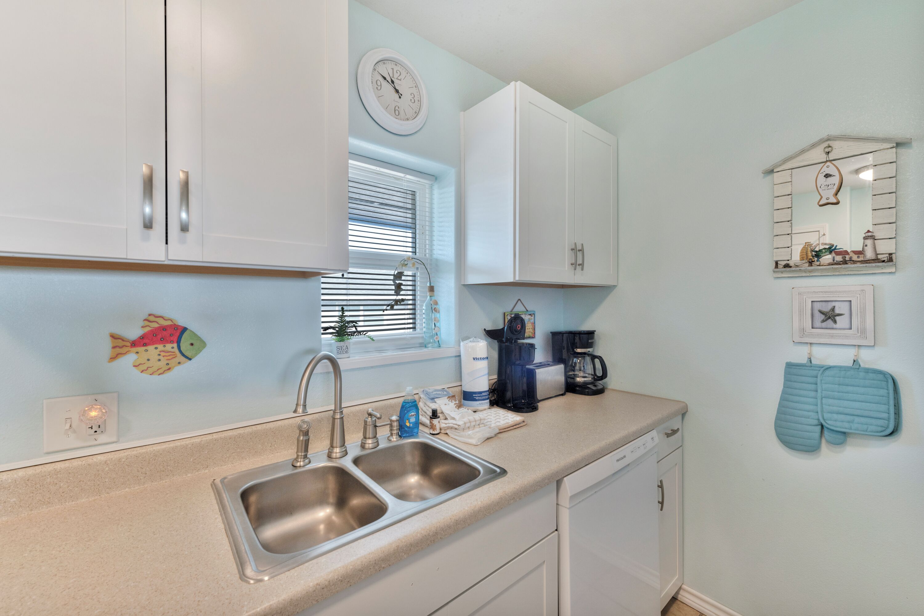 A compact kitchen with a double sink, white cabinets, a clock, and a small window with blinds.