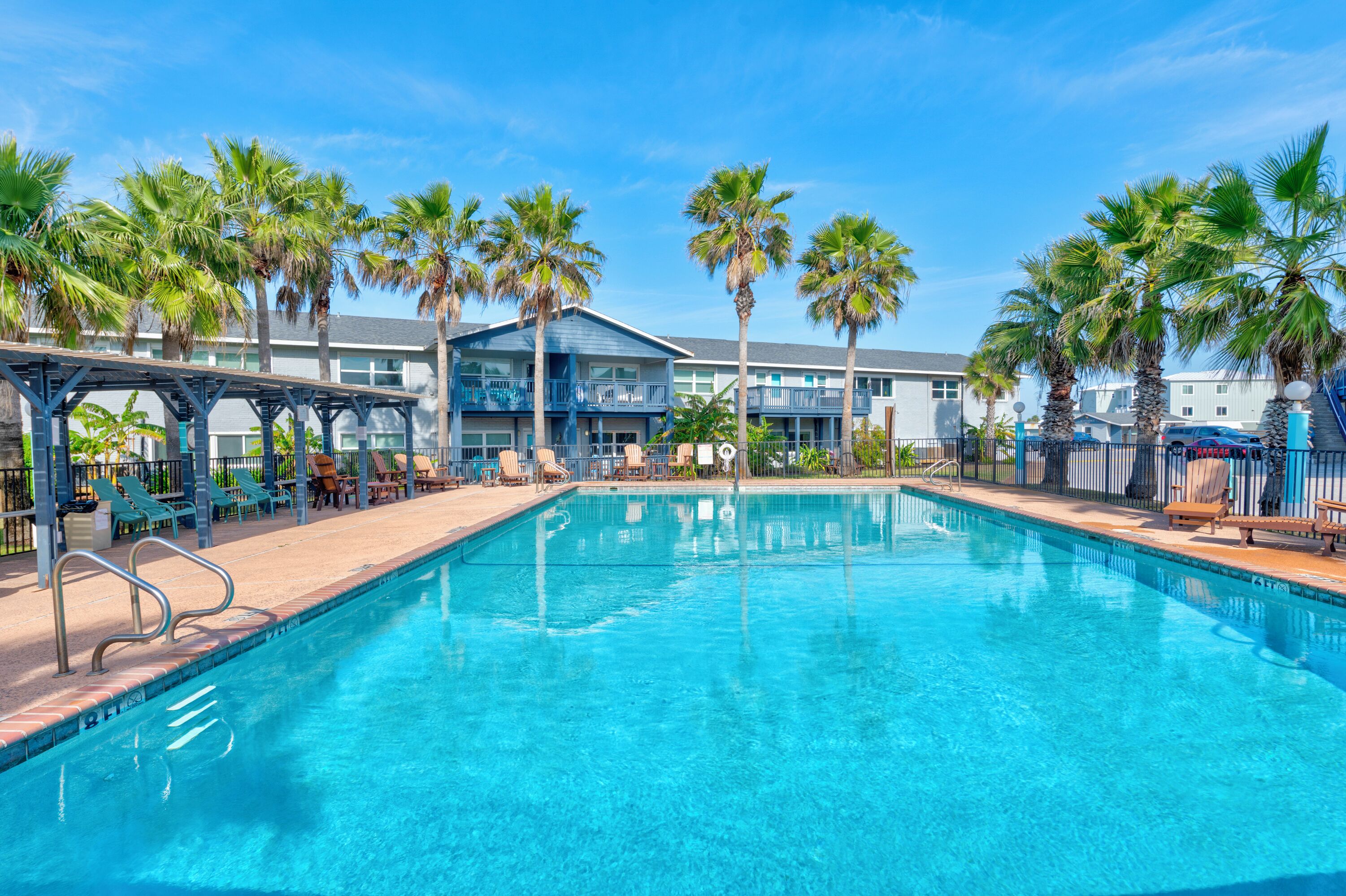 A clear blue pool with lounge chairs and palm trees, surrounded by a building with multiple balconies.