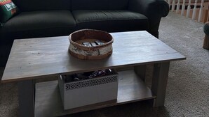 Una sala de estar acogedora con un sofá verde oscuro, una mesa de centro de madera con una cesta, y vistas al exterior a través de grandes ventanales.