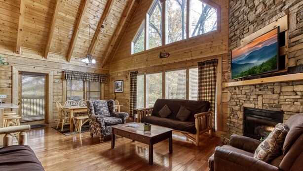Interior acogedor de una cabaña con paredes de madera, chimenea de piedra, televisor montado encima y una zona de comedor con mesa y sillas.