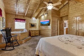 A log cabin bedroom with a wooden ceiling, a ceiling fan, a large bed, a rocking chair, a bathroom with a sink and mirror, and a TV mounted on the wall.