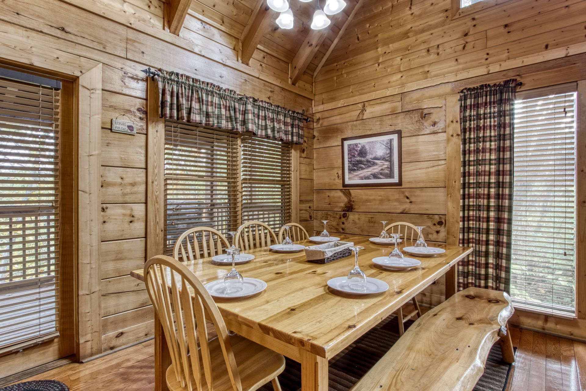 A wooden dining room with a table set for six, flanked by two windows with blinds, a framed picture on the wall, and a sign indicating the room is for sale.
