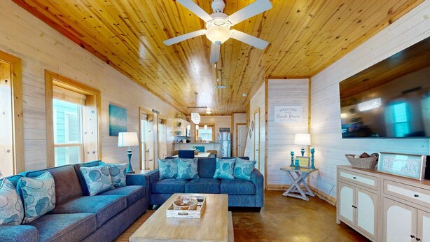 A cozy cabin interior with a wooden ceiling, a large sofa, a coffee table, and a kitchen area. - Townhome Located in Town, Close to beach, Shared Pool, Pet Friendly (Port Aransas)