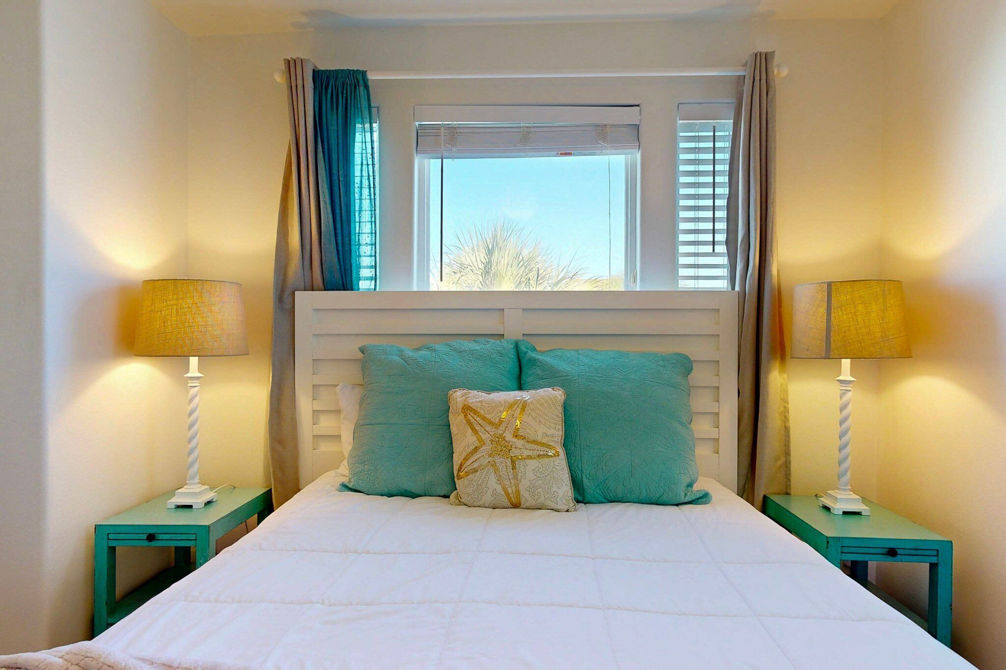 A neatly made bed with a white comforter and teal pillows, flanked by two bedside tables with lamps, in a room with a window and curtains.