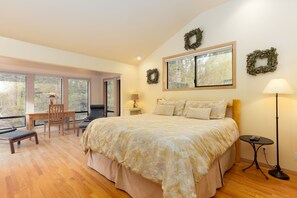 A bedroom with a large bed, a desk, and a chair. There are two windows and a view of trees.