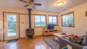 A spacious living room with a fireplace, a ceiling fan, and large windows offering a view of the outdoors. - Family vacation destination! Lake activities and game house! (Kenai)