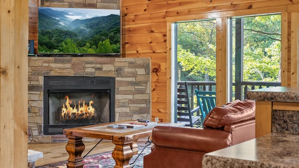 Una sala de estar acogedora con chimenea de piedra, un televisor montado en la pared y vistas a un bosque a través de amplios ventanales.