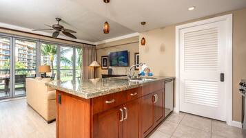 A modern kitchen with a granite countertop, wooden cabinets, and a large island. There is a ceiling fan, pendant lights, and a view of a balcony with a palm tree.