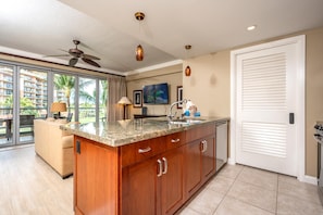 A modern kitchen with a granite countertop, wooden cabinets, and a large island. There is a ceiling fan, pendant lights, and a view of a balcony with a palm tree. - Honua Kai | Ocean View 1 BR | Family Friendly | Car Incl. w/ 6+ Nights | HKH-344 by KBM (Lahaina)