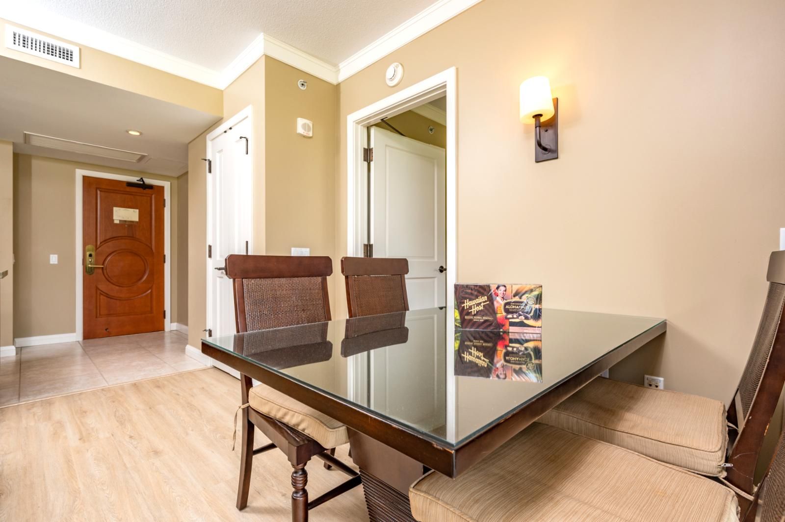 A hotel room with a glass dining table, two brown chairs, a wall-mounted light, and a poster on the wall.