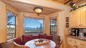 A cozy dining area with a round wooden table and red cushioned chairs, large windows with a view of a forest and mountains, and wooden cabinetry in the adjacent kitchen area.