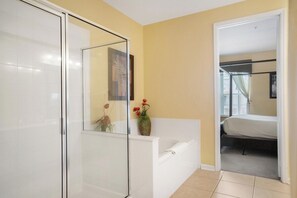 A bathroom with a glass shower enclosure, a white bathtub, and a door leading to a bedroom.