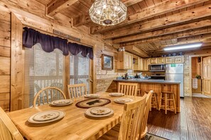 L’intérieur d’un chalet en bois comprend une table à manger pour quatre personnes, des armoires en bois, un coin cuisine équipé d’appareils électroménagers et un lustre.