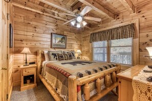 Une chambre dans une cabane en bois, comprenant un lit, une table de chevet, un bureau, un ventilateur de plafond et une fenêtre avec des stores.